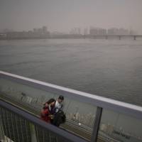 With Seoul’s skyline behind them, a young couple are reflected in the glass of a barrier as they walk across a bridge over the Han River when the capital was shrouded by high levels of Chinese sand and PM 2.5-laden smog Monday. High levels of yellow sand, PM2.5 heading in from China