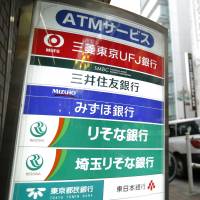A pedestrian walks past a board displaying signage for banks including Bank of Tokyo Mitsubishi UFJ, Sumitomo Mitsui Banking Corp. and Mizuho Bank Ltd. in Tokyo in May 2015. Clouds lifting for Japan's banks as negative rates stay put