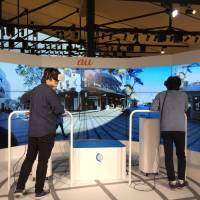 People try out KDDI’s virtual traveling service on Nov. 2 in Tokyo. Wearing headsets, the two people are vicariously exploring Manly Beach, Australia, in real time, seeing what a travel agent sees on the screen around them. VR wave to leave Japan awash in business opportunities