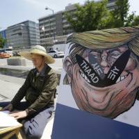 A South Korean protester sits next to a poster with an illustration of U.S. President Donald Trump to oppose a plan to deploy an advanced U.S. missile defense system called Terminal High-Altitude Area Defense near the U.S. Embassy in Seoul on Monday. South Korea to freeze deployment of THAAD missile defense pending environmental probe