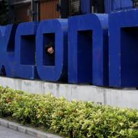 A worker looks out through the logo at the entrance of the Foxconn complex in the southern Chinese city of Shenzhen in 2010. Taiwanese electronics maker Foxconn’s plan to build a display panel factory in the U.S. has sparked a flurry of lobbying by states vying to land what some economic development officials say is a once-in-a-generation prize. Rust Belt and other U.S. areas seen vying for big Foxconn display panel factory