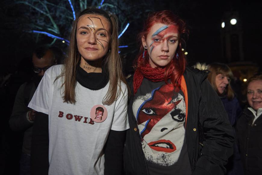 Two young fans in the crowd wear Bowie merchandise and Ziggy Stardust face makeup in his honor.