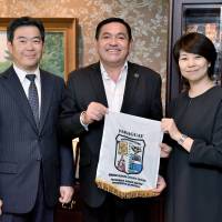 Teodosio Romilio Gomez Ibañez (center), mayor of the city of  Villeta, Paraguay,  visits The Japan Times\' Tokyo office on Oct. 10, and hands a pennant bearing the city\'s coat of arms to Chairperson and Publisher Minako Kambara Suematsu. On the left is Japan Times President Takeharu Tsutsumi. | YOSHIAKI MIURA