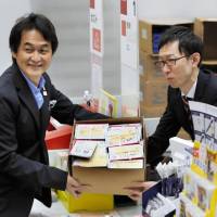 Takeshi Natsuno (left), a member of the panel choosing the mascot for the Tokyo Olympics, displays a box of postcards Monday with information on the voting process to be sent to elementary schools nationwide. | POOL / VIA KYODO