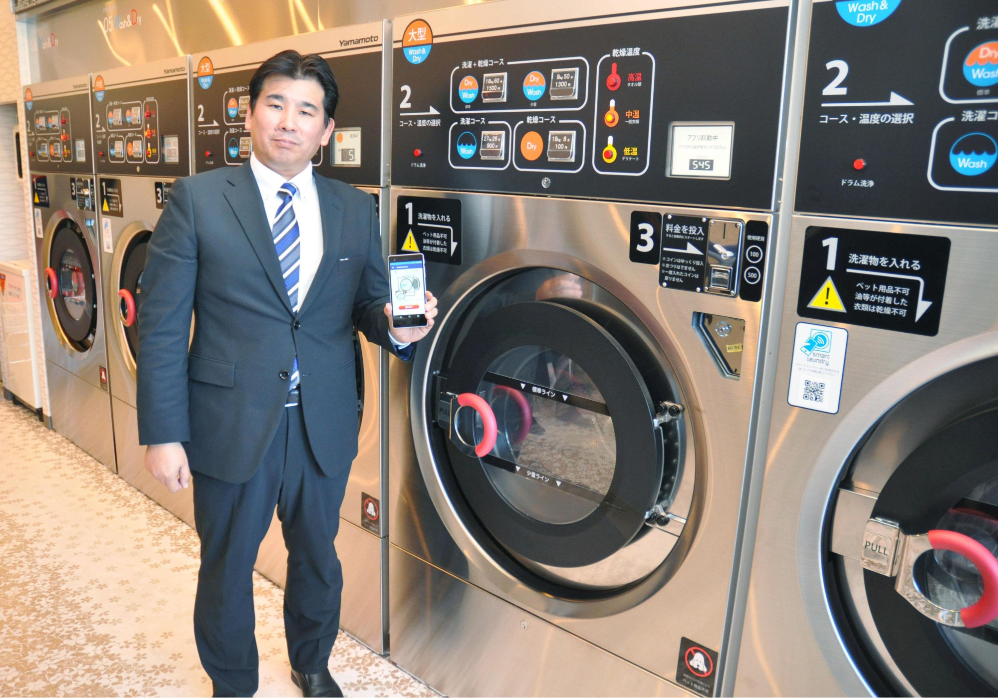 Wash Plus President Kentaro Takanashi poses with a smartphone at one of his laundromats in the city of Chiba. His company's smartphone service lets customers check whether washing machines are available, their clothes are ready or make payments. | KYODO