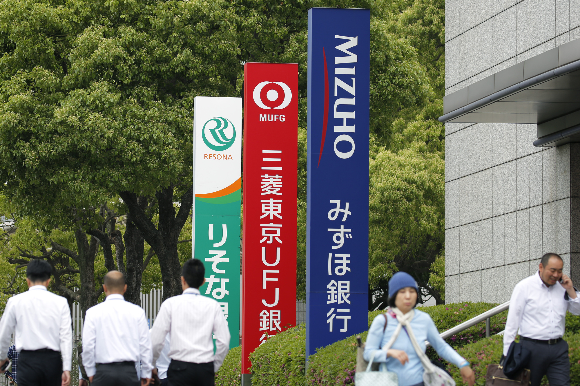 Pedestrians walk past signs for Resona Bank Ltd., Bank of Tokyo Mitsubishi UFJ Ltd. and Mizuho Bank Ltd. in Tokyo in May 2015. | BLOOMBERG