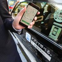 A representative of Nihon Kotsu Co. demonstrates a ride-sharing app beside a taxi in Tokyo\'s Chiyoda Ward on Wednesday. | YOSHIAKI MIURA