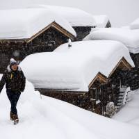 A person walks in front of chalets covered with fresh snow, in Bellwald, Switzerland, on Sunday. Heavy snowfall increased the risk of avalanches in almost all the Swiss mountains. | JEAN-CHRISTOPHE BOTT / KEYSTONE / VIA AP