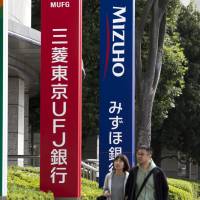 Pedestrians walk past bank signboards in central Tokyo. The industry is at a crossroads as the central bank maintains an ultraeasy monetary policy and new technology sees growing prominence in the sector. | BLOOMBERG