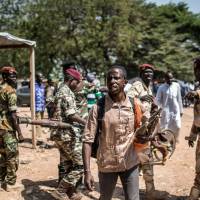 A member of the Muslim auto defense group of the flashpoint PK5 district in Bangui carries RPG ammunitions as he walks by Central African Armed forces soldiers (FACA &#8212; Forces Armees de Centre Afrique) in Bangui to secure the voting operations at the Koudoukou school where earlier a splinter group disrupted the delivery of the electoral material in 2015. U.N. and Cntral African security forces have launched an operation against the Muslim auto-defense group, leaving at least 30 people wounded, including two U.N. peacekeepers. | AFP-JIJI