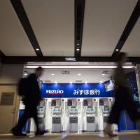 People walk past a Mizuho Bank ATM in Tokyo. Japan\'s three megabanks including Mizuho are considering jointly managing and developing ATMs as part of cost-cutting efforts. | BLOOMBERG