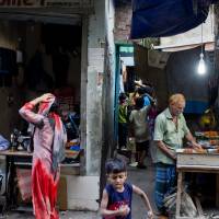 Residents walk past a man making barbecue in an alley at the Mohammadpur Geneva camp, where over 100 suspected drug peddlers were detained in a raid by Rapid Action Battalion (RAB) in Dhaka. | AP