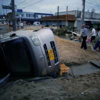 Local residents walk next to a damaged car in a flooded area of Kurashiki, Okayama Prefecture, on Sunday. | REUTERS