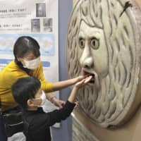Visitors to Osaka University Hospital put their hands into a sanitizer dispenser that is shaped like the famous Roman sculpture the \"Mouth of Truth\" on Tuesday. | KYODO