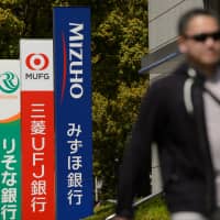 A pedestrian walks past signs for banks in Tokyo. Two megabanks, MUFG Bank and Sumitomo Mitsui Banking Corp., plan to share their respective automated teller machines to cut operational costs, according to sources. | BLOOMBERG