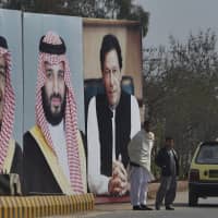 A man with his son waits for transport next to huge portraits of Saudi leaders and the Pakistani prime minister displayed in Islamabad on Sunday on the occasion of the visit by Saudi Arabia\'s crown prince. | AP