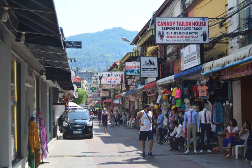 Tourists walk down a street in Patong featuring a number of tailoring shops.