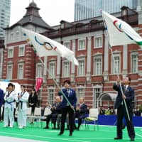 Tokyo Gov. Yuriko Koike and Yoshitaka Sakurada, minister for the Tokyo Olympic and Paralympic Games, wave the official Olympic flags in front of Tokyo Station on Saturday. | RYUSEI TAKAHASHI