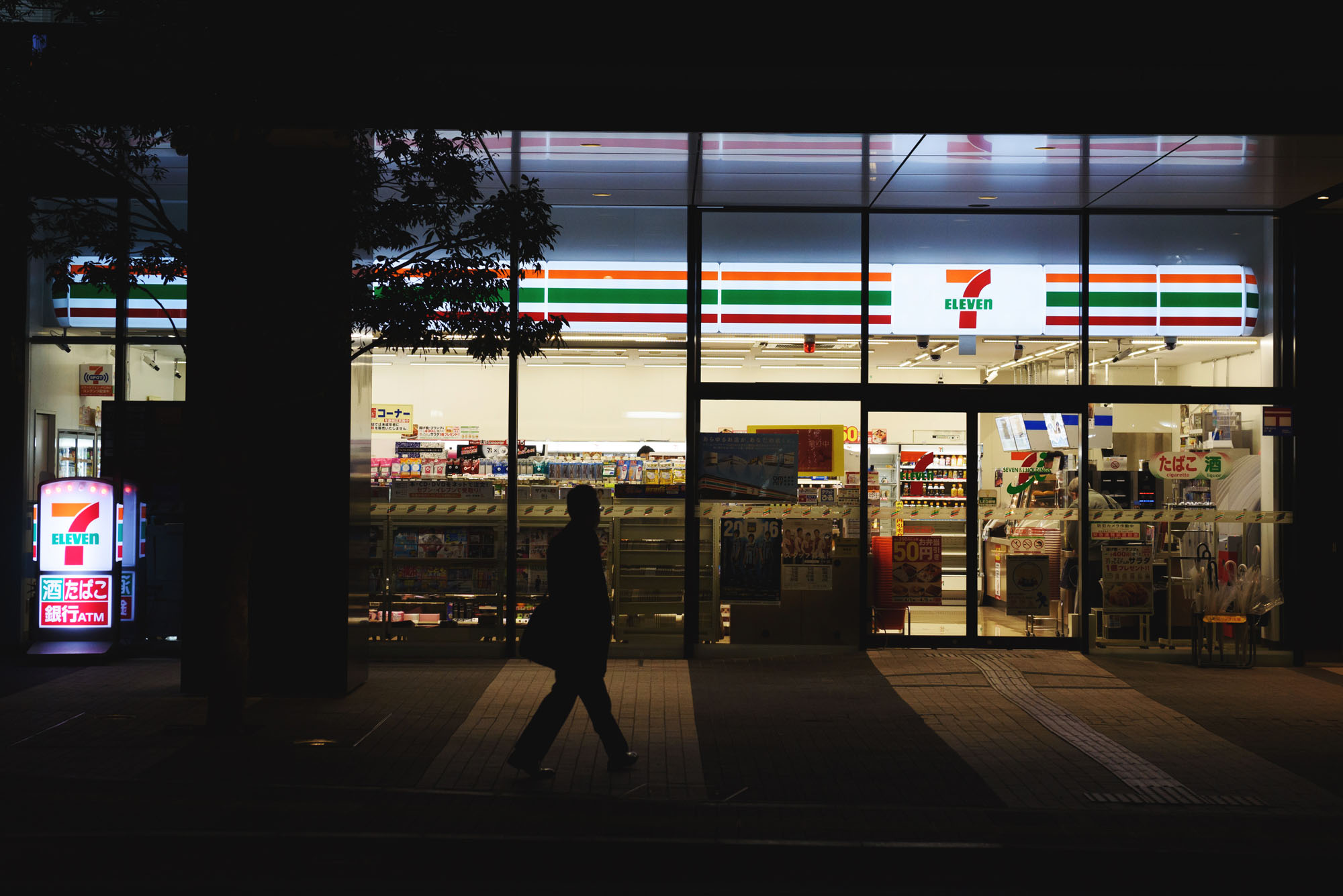 A pedestrian walks past a 7-Eleven convenience store, operated by Seven &amp; I Holdings Co., at night in Kawasaki in April 2016. | BLOOMBERG