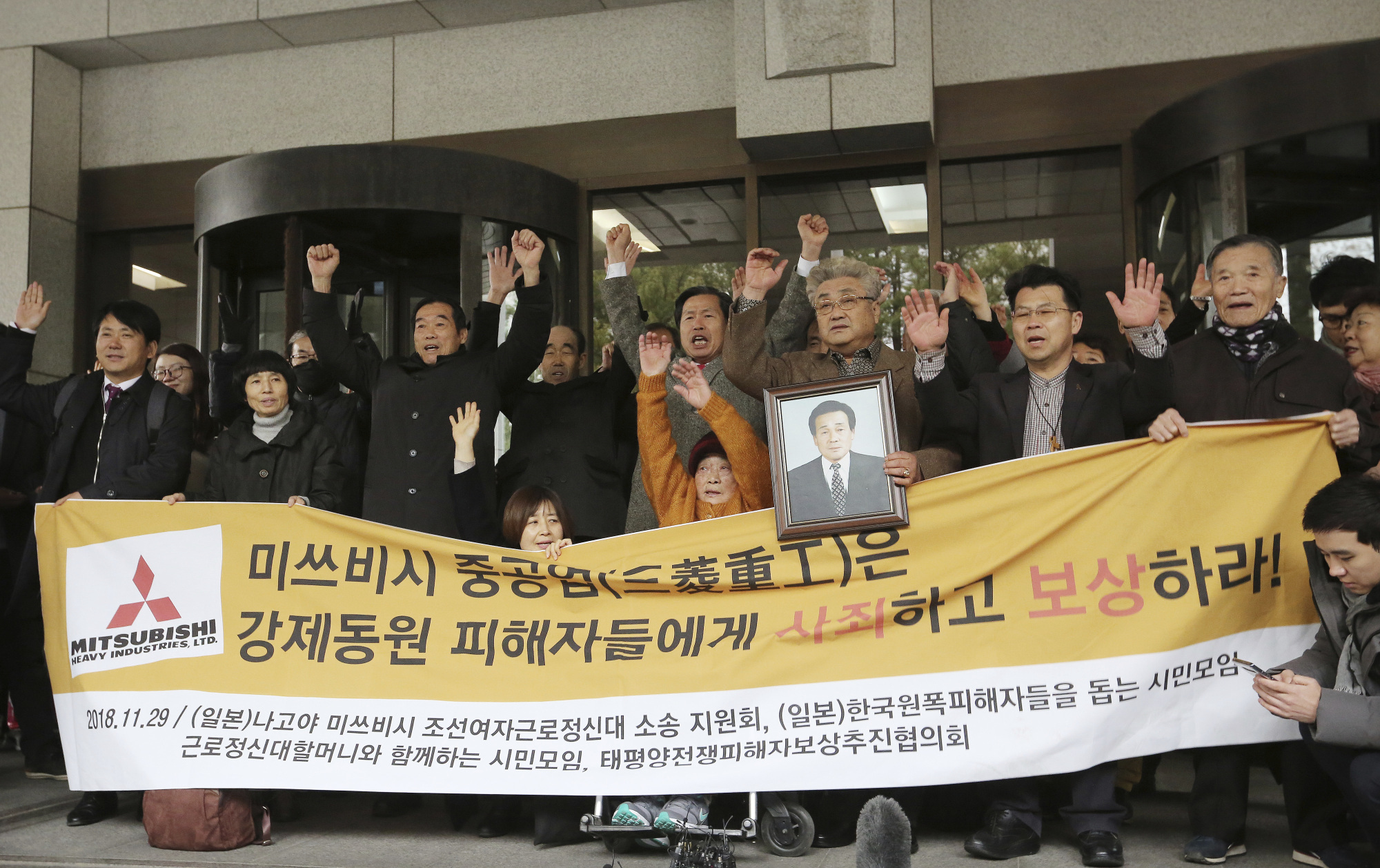 Korean victims of Japan's forced wartime labor raise hands with family members in Seoul on Nov. 29, 2018, after the Supreme Court ordered Mitsubishi Heavy Industries to compensate them. Seoul has accused Tokyo of weaponizing trade to retaliate against similar court rulings calling for Japanese companies to offer reparations to Koreans who were forced into slave labor during World War II. The two nations, however, settled war-related compensation in the 1965 pact that normalized bilateral relations. The signs says: 'Mitsubishi Heavy Industries apologizes and compensates to victims.' | AP