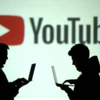 Silhouettes of mobile device users are seen next to a screen projection of the Youtube logo in 2018. | REUTERS