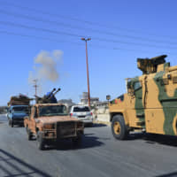 Smoke rises in the background from a bombardment around the area as fighters with the Free Syrian army drive their pickup truck past a Turkey Armed Forces convoy, at a highway between Maaret al-Numan and Khan Sheikhoun in Idlib province, Syria, Monday. | MURAT KIBRITOGLU / DHA / VIA AP