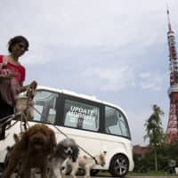 A woman walks with dogs in front of the Navya Technologies SAS Arma autonomous shuttle bus during a media preview of a test drive in Tokyo in July 2017. | BLOOMBERG