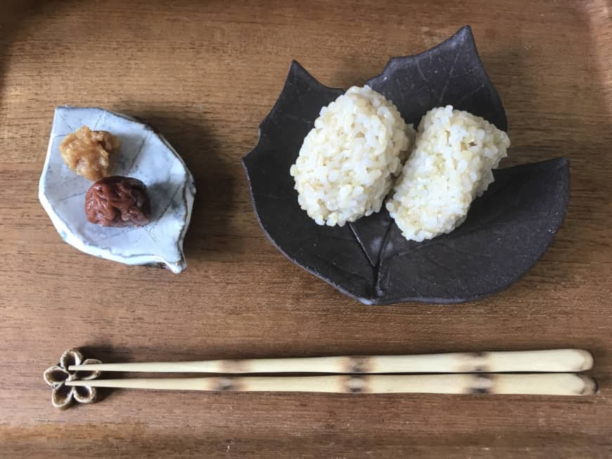 'I will have this meal': A pair of chopsticks sit horizontally in front of brown rice onigiri (rice balls) and a homemade pickled plum with miso. | MOMOKO NAKAMURA
