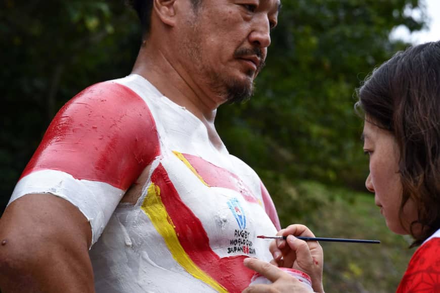 Hiroshi Moriyama has his torse painted by his wife, Rika, in the colors of Japan's Brave Blossoms near Shizuoka Stadium Ecopa Stadium on Saturday. | AFP-JIJI