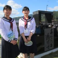 Yukime Sasaki (right) and another high school student pose for a photo next to the prayer monument installed at Kamaishi Unosumai Recovery Memorial Stadium before Wednesday\'s Uruguay-Fiji game. They serve as memory keepers of the 2011 disaster. | KAZ NAGATSUKA