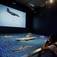 Visitors watch a TV screen showing a U.S. Air Force F-35 fighter jet firing a missile near models of China\'s Liaoning aircraft carrier with navy frigates and submarines on display at a military museum in Beijing on Aug. 1. | AP