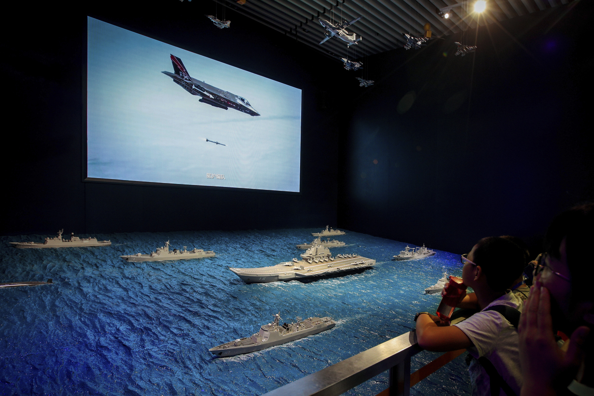 Visitors watch a TV screen showing a U.S. Air Force F-35 fighter jet firing a missile near models of China's Liaoning aircraft carrier with navy frigates and submarines on display at a military museum in Beijing on Aug. 1. | AP