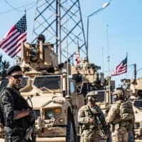 A member of the Kurdish People\'s Protection Units (left) stands guard as U.S. military armored vehicles and soldiers patrol near an oil well in Rumaylan (Rmeilan) in Syria\'s northeastern Hasakeh province on Wednesday. | AFP-JIJI