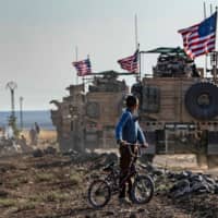 A Syrian boy on his bicycle looks at a convoy of U.S. armored vehicles patrolling fields near the northeastern Syria town of Qahtaniyah at the border with Turkey Thursday. U.S. forces accompanied by Kurdish fighters of the Syrian Democratic Forces (SDF) patrolled part of Syria\'s border with Turkey, in the first such move since Washington withdrew troops from the area earlier in October, an AFP correspondent reported. | AFP-JIJI