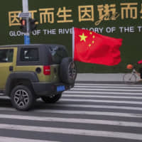 A vehicle flying a Chinese flag drives past a woman wearing a face mask as she rides a bicycle along a street in Wuhan in central China\'s Hubei Province Wednesday. Countries began evacuating their citizens Wednesday from the Chinese city hardest-hit by a new virus that has now infected more people in China than were sickened in the country by SARS. | AP
