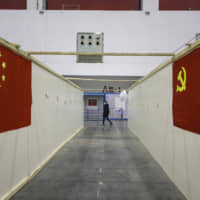 A person wearing a mask walks past a corridor withe a Chinese national flag (left) and a Chinese Communist Party flag adorn walls in a temporary hospital converted from an exhibition center in Wuhan, China, on Feb. 18. Beijing formally decided Monday to postpone the annual meeting of the country\'s national parliament. | AP