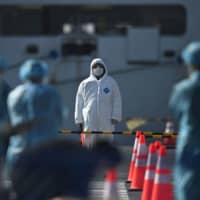 Workers in protective clothes stand before passengers disembarkating from the Diamond Princess cruise ship, in quarantine due to fears over the new COVID-19 coronavirus, at Daikoku Pier Cruise Terminal in Yokohama on Friday. | AFP-JIJI