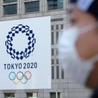 Let the posiivity begin: A man wearing a face mask passes the logo of the Tokyo 2020 Olympic Games displayed on the Tokyo Metropolitan Government Building in March. | AFP-JIJI