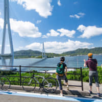 A pair of cyclists take a break near the Tatara Bridge on the Shimanami Kaido expressway in Onomichi, Hiroshima Prefecture. | CITY OF ONOMICHI