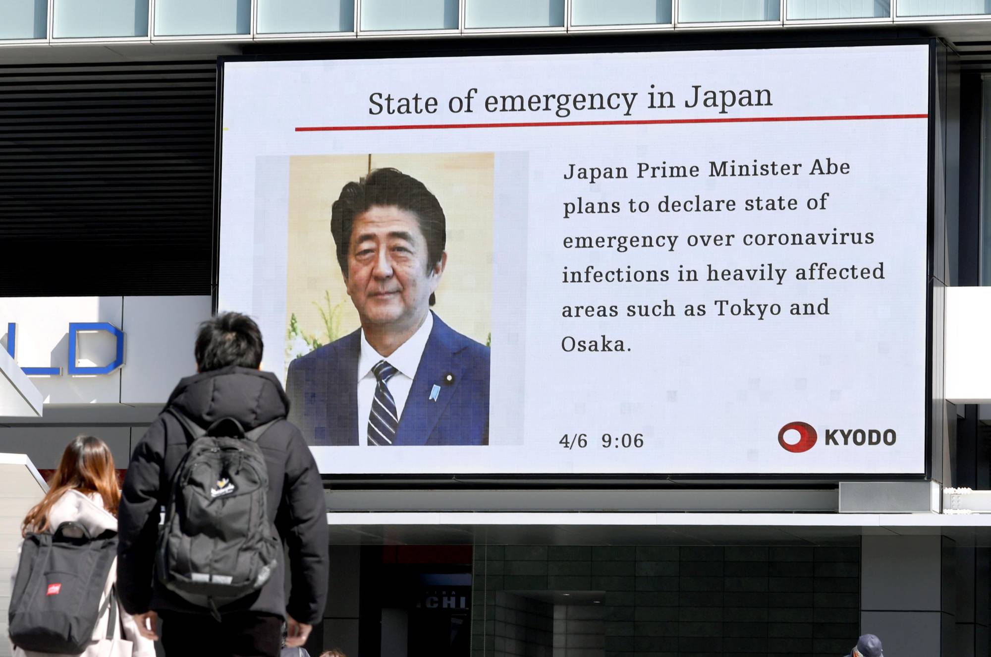 People walk by a big screen in the Akihabara district of Tokyo on Monday broadcasting news that Prime Minister Shinzo Abe is set to declare a state of emergency over the growing spread of COVID-19 in Japan. | KYODO