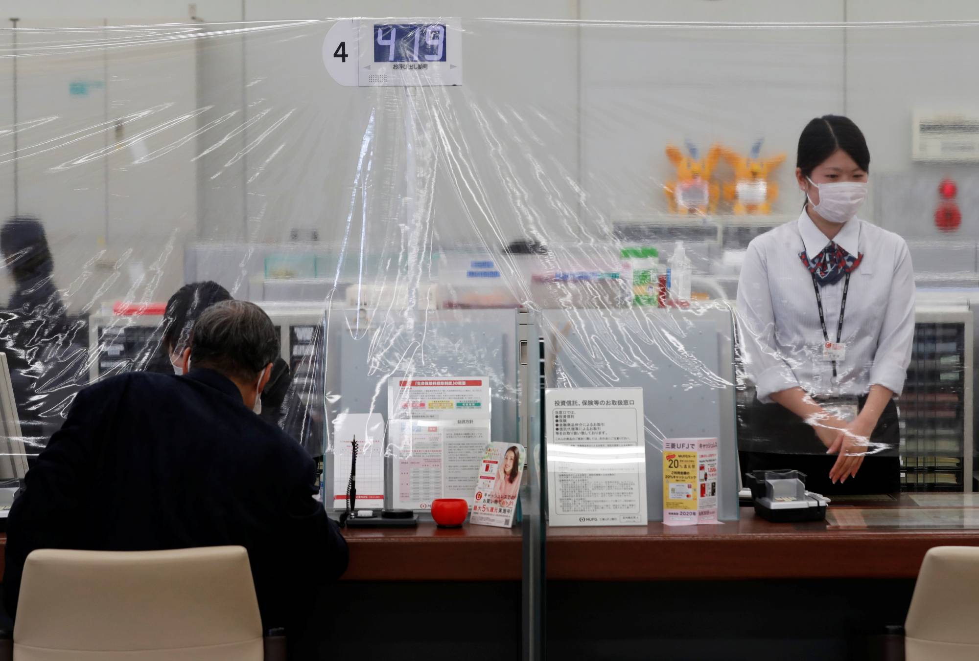 An employee at a MUFG Bank branch in central Tokyo on Friday stands behind a plastic curtain installed to prevent coronavirus infections. | REUTERS