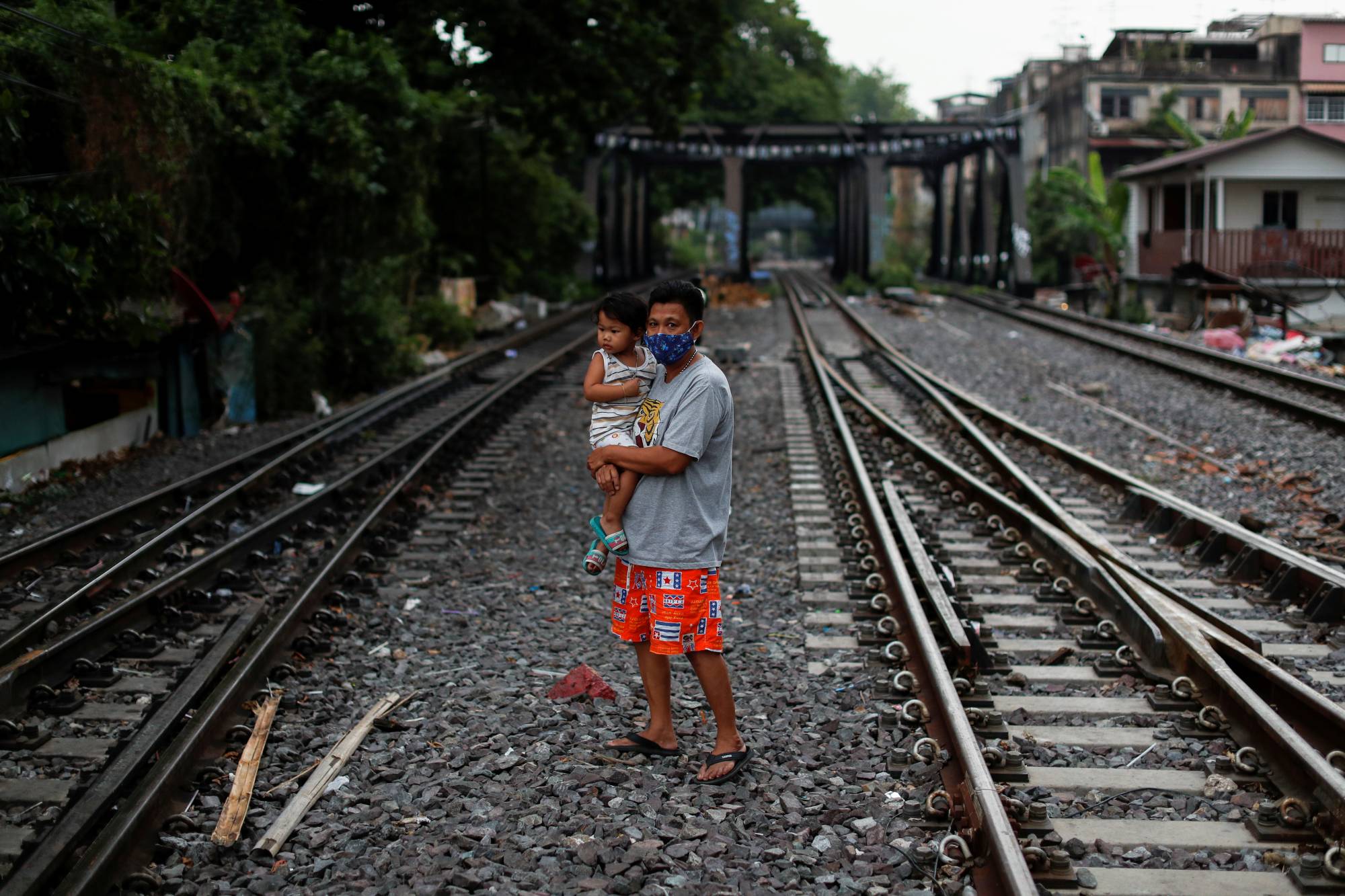 A man wearing a protective mask holds a child as he crosses the tracks in a slum in Bangkok on Monday. | REUTERS