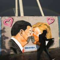 A man walks in front of graffiti, displayed at a wall in the public park Mauerpark in Berlin, depicting U.S. President Donald Trump and China\'s President Xi Jinping kissing each other with face masks on Wednesday. | AP
