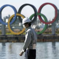 A masked construction worker stands in Tokyo\'s Odaiba waterfront district, with the Olympic rings in the background.
 | AP