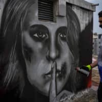 Sixteen-year-old Greek graffiti artist S.F. paints on the roof of a building in Athens last month. The image depicts a woman with injuries on her face and her finger over her lips, inspired by the increase in cases of domestic violence amid worldwide lockdowns. | AFP-JIJI