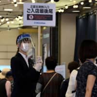 An employee wearing a face shield holds up a sign as people wait in a queue to visit a Matsuzakaya department store in Tokyo as it reopened for business on May 26. | AFP-JIJI