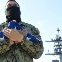 Aviation Ordnanceman Airman Andrew Halford holds the American flag on the flight deck of the aircraft carrier USS Theodore Roosevelt as it departs following an extended visit  to Apra Harbor, Guam, due to a coronavirus outbreak, on May 21. | U.S. NAVY / VIA REUTERS