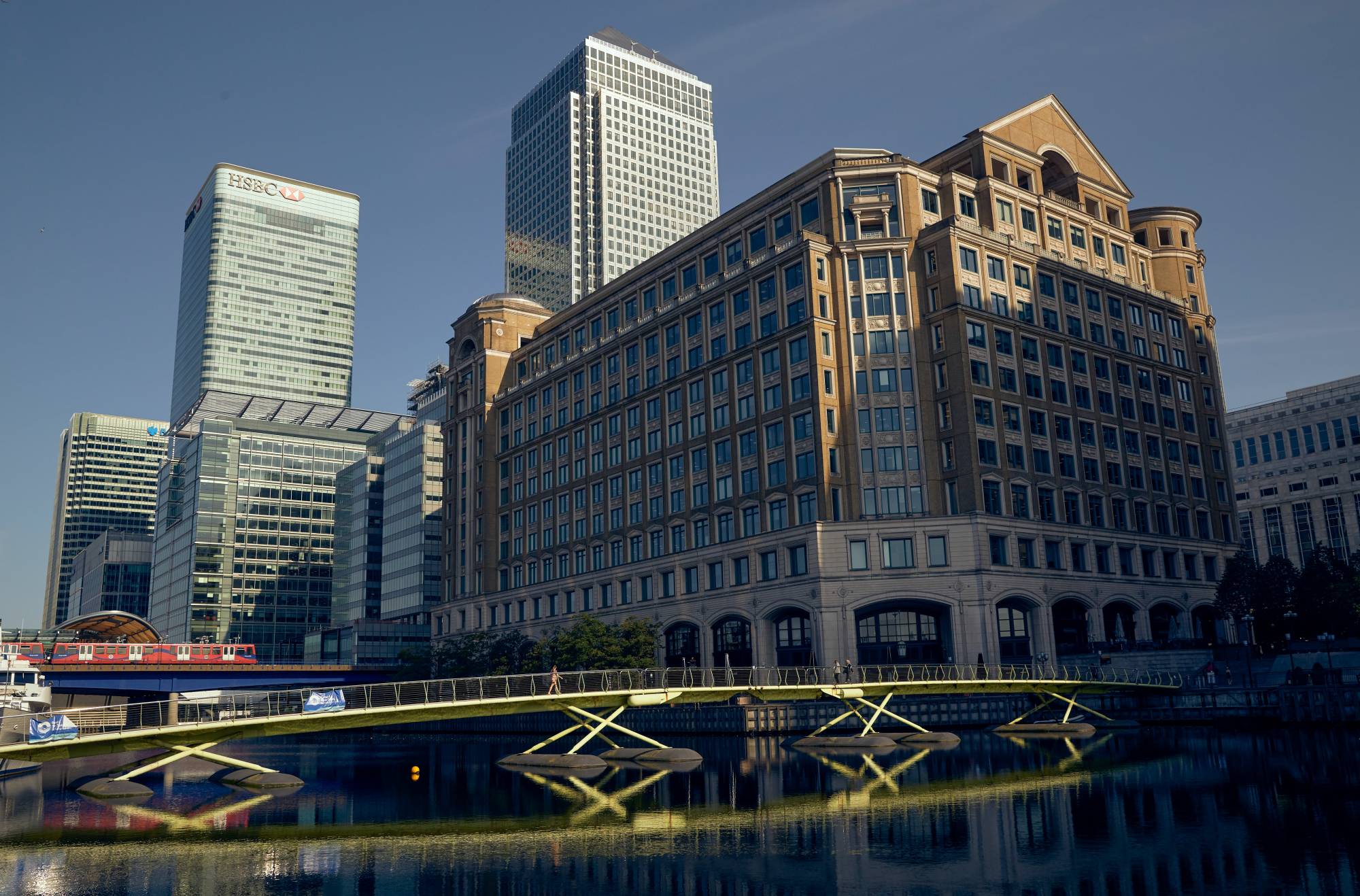 Pedestrians cross a footbridge in view of buildings in the West India Dock in the Canary Wharf financial district in London. | Bloomberg