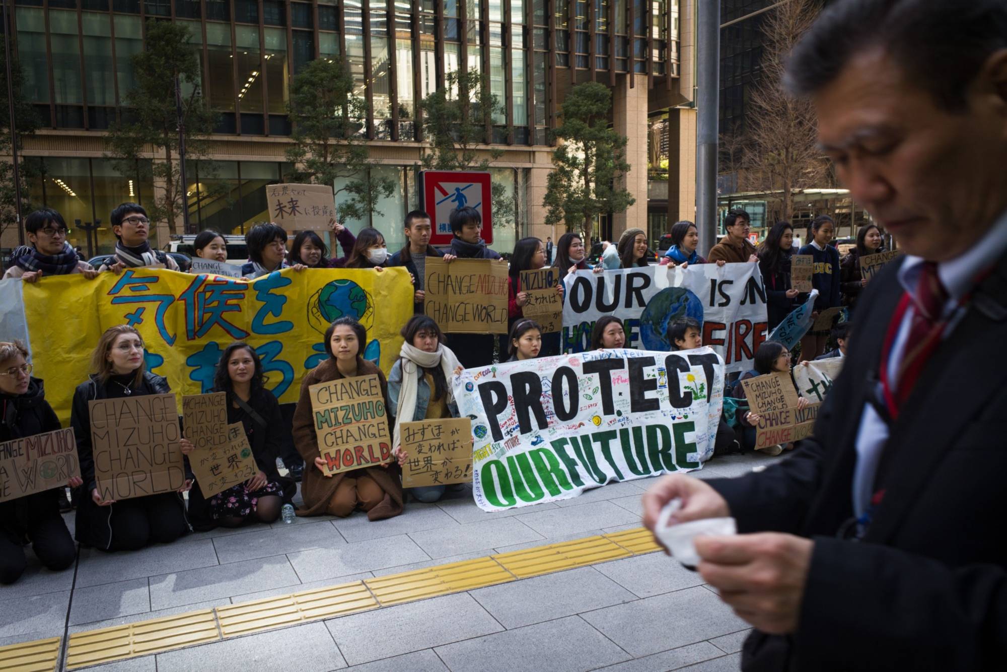 A pedestrian walks past a group of youth climate activists demonstrating outside the Mizuho Financial Group Inc. headquarters in Tokyo in March. The anti-coal demonstration organized by Fridays for Future Tokyo denounced Mizuho Bank's role in financing coal projects globally. | BLOOMBERG