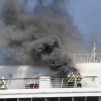 Smoke rises from the Japanese cruise ship Asuka II, docked in Yokohama Port, on June 16. Finding places to park idle ships is just the first painful point. To keep things shipshape and avoid costly repairs and other issues, the vessels must also be kept operating. | AP